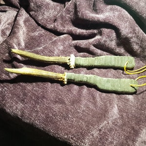 Puede incluir: Dos herramientas con mango de asta de ciervo con envolturas de cuero verde oscuro y cordones amarillos. Las herramientas están sobre una superficie de tela de color morado oscuro.