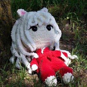 Puede incluir: Una muñeca de crochet blanca con el pelo largo blanco y un vestido rojo está tumbada en el suelo cerca de un árbol.