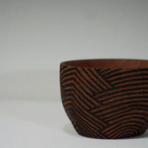 May include: A handcrafted wooden bowl with a dark brown finish and a geometric pattern of black lines. The bowl has a rounded shape and is suitable for serving or display.