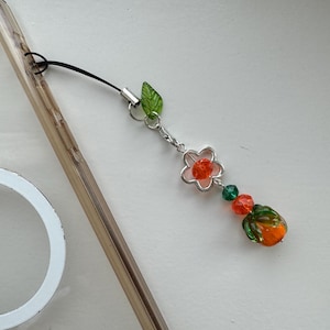 May include: A phone charm with a silver chain, a green leaf, a pink flower with an orange center, green beads, and an orange and green bead in the shape of a fruit.