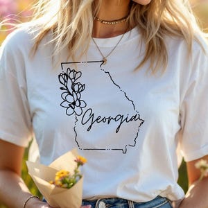 May include: White t-shirt featuring a black outline of the state of Georgia with the word "Georgia" in script and a floral design. The shirt is made of a soft material and is suitable for casual wear.