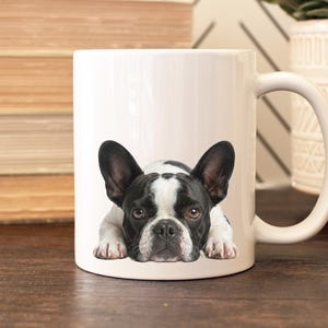 Op de afbeelding: Witte keramische mok met een zwart-wit Frans Bulldog ontwerp. De hond wordt liggend afgebeeld. De mok heeft een gebogen handvat en staat op een houten oppervlak.