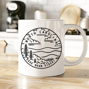 May include: White ceramic mug with a black outline of a circle containing a landscape scene with mountains, trees, and clouds. The text "NORTH CAROLINA" is written around the circle. The text "ESSE QUAM VIDERI" is written below the circle.