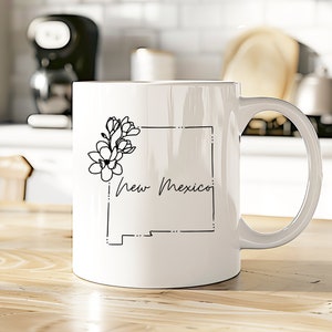 May include: White ceramic coffee mug with a black outline of the state of New Mexico and a floral design. The text "New Mexico" is written in cursive.