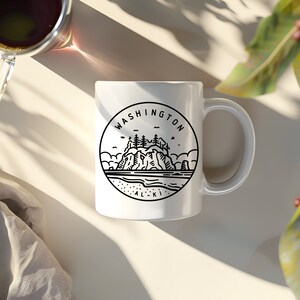 May include: White ceramic mug with a black line drawing of a coastal scene, including the text "WASHINGTON ALKI" in a circular design. The mug has a curved handle and is set on a light-colored surface.