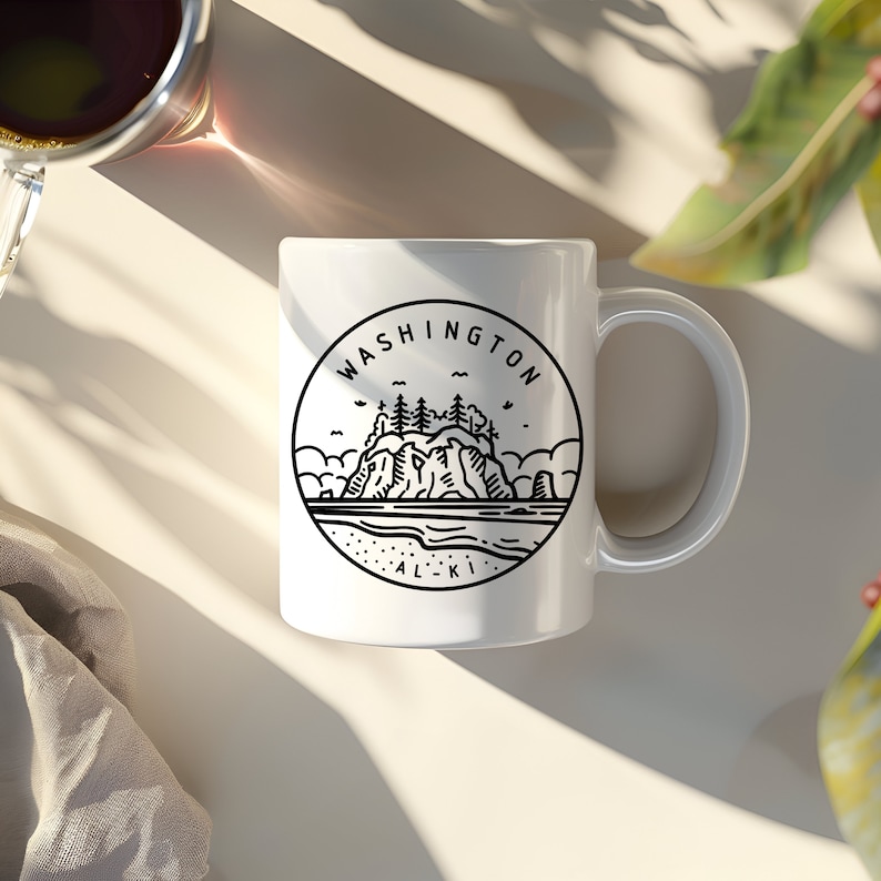 May include: White ceramic mug with a black line drawing of a coastal scene, including the text "WASHINGTON ALKI" in a circular design. The mug has a curved handle and is set on a light-colored surface.