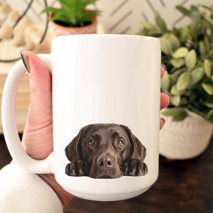 May include: A white ceramic mug featuring a detailed image of a chocolate Labrador Retriever's face. The dog's head and paws are at the bottom of the mug. The mug has a large handle.