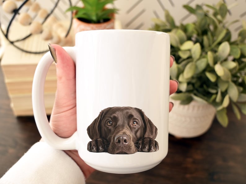 May include: A white ceramic mug featuring a detailed image of a chocolate Labrador Retriever's face. The dog's head and paws are at the bottom of the mug. The mug has a large handle.