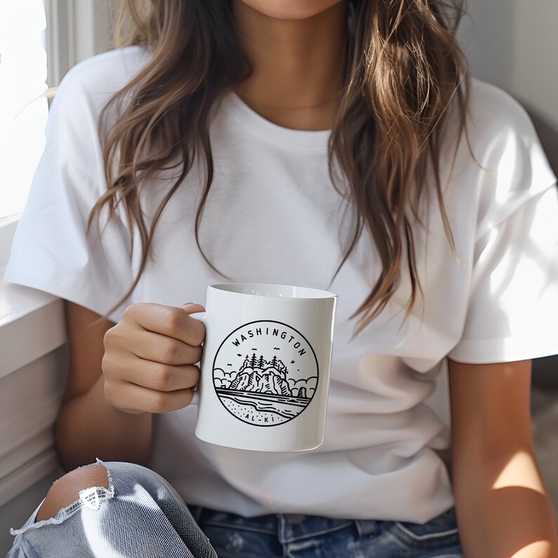 May include: White ceramic mug featuring a black and white illustration of a coastal landscape with the text "WASHINGTON ALKI" in a circular design. The mug has a handle and is held by a person wearing a white t-shirt.