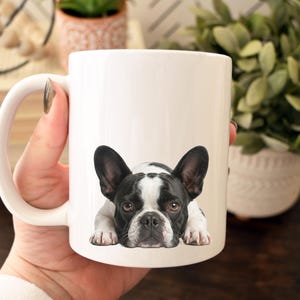 Op de afbeelding: Witte keramische mok met een zwart-wit Franse Bulldog ontwerp. De hond wordt liggend afgebeeld. De mok heeft een gebogen handvat. De mok wordt door een persoon vastgehouden.