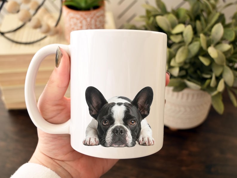 Op de afbeelding: Witte keramische mok met een zwart-wit Franse Bulldog ontwerp. De hond wordt liggend afgebeeld. De mok heeft een gebogen handvat. De mok wordt door een persoon vastgehouden.