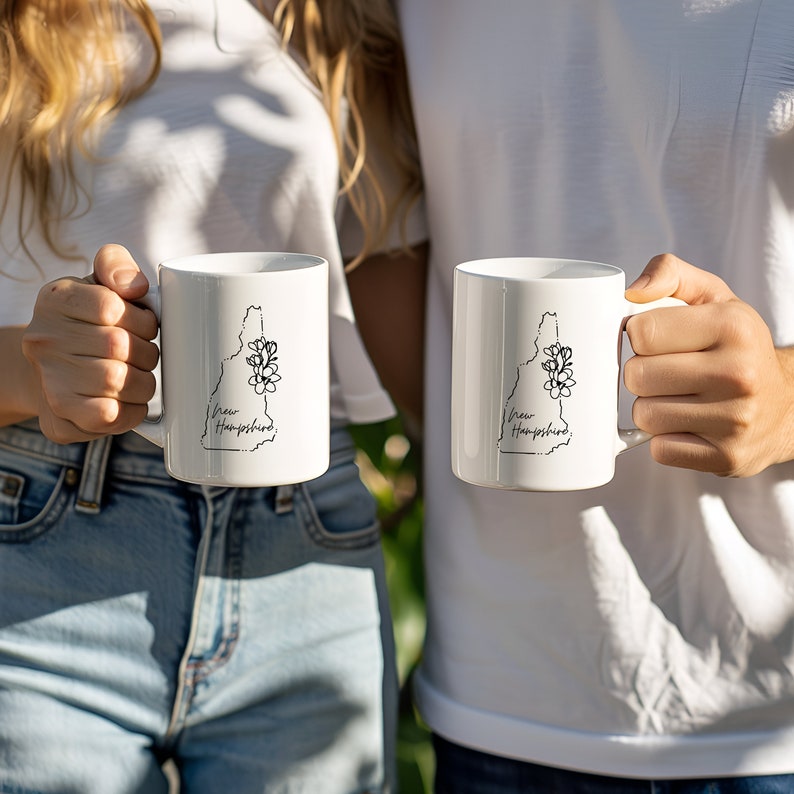 May include: Two white ceramic mugs with black outlines of the state of New Hampshire and the words "New Hampshire" printed on them.