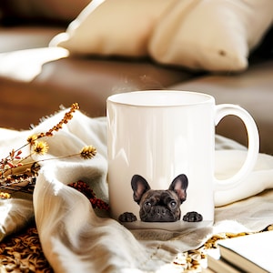 May include: White ceramic mug with a black and brown French bulldog peeking over the rim. The dog's face is visible, with its tongue sticking out slightly. The mug is sitting on a light brown surface.