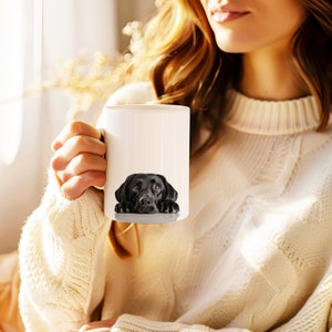 May include: A white ceramic mug with a black Labrador Retriever peeking over the rim. The dog's head and paws are visible, with a white background behind it.