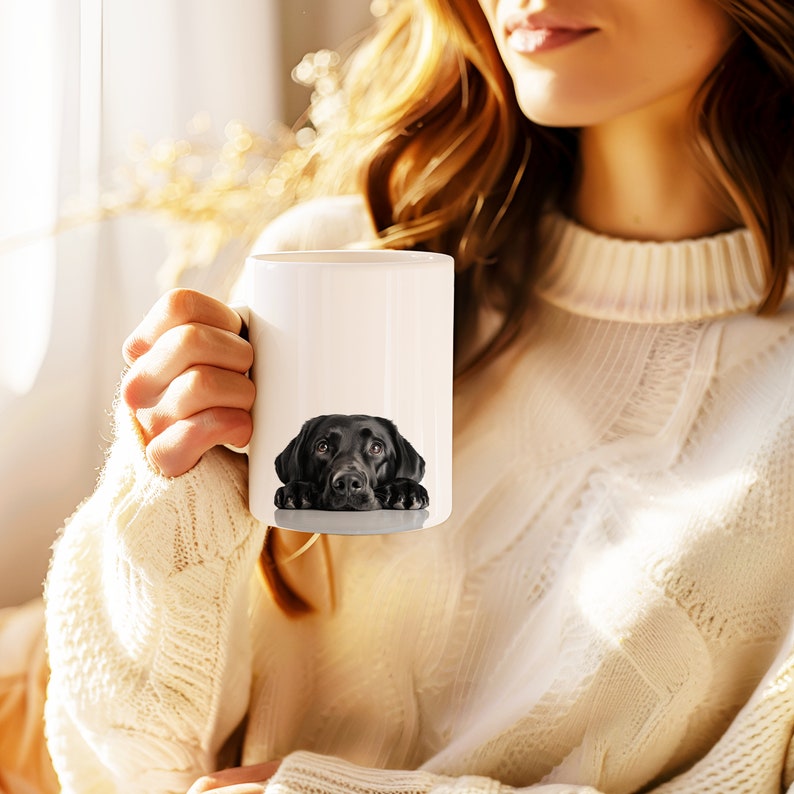May include: A white ceramic mug with a black Labrador Retriever peeking over the rim. The dog's head and paws are visible, with a white background behind it.