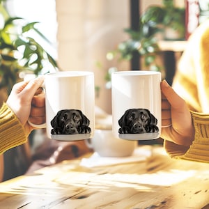 May include: Two white ceramic mugs with black labrador retriever dog faces printed on them. The dogs are looking at the camera with their heads resting on the rim of the mugs.