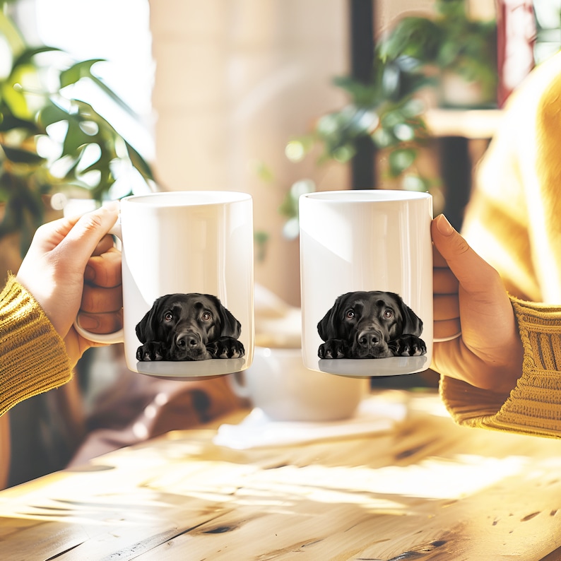 May include: Two white ceramic mugs with black labrador retriever dog faces printed on them. The dogs are looking at the camera with their heads resting on the rim of the mugs.