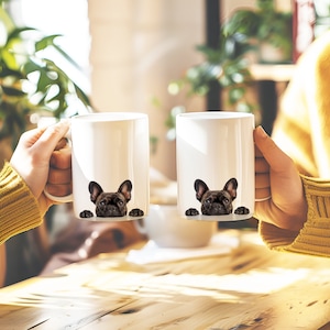 May include: Two white ceramic mugs with a black French bulldog peeking over the rim of each mug. The mugs are held by hands in a close-up shot.