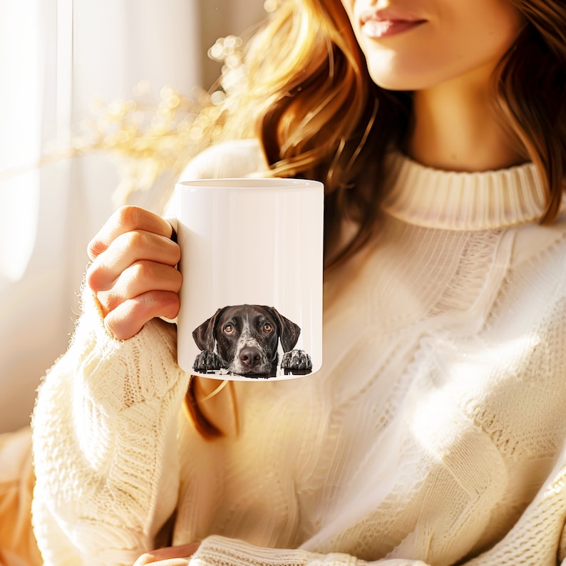 May include: A white ceramic mug with a black and white dog peeking over the rim. The dog has brown eyes and a black nose.