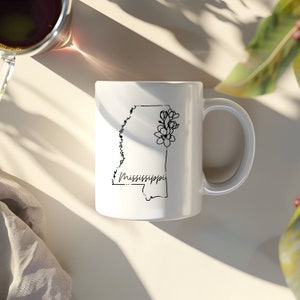 Pode incluir: Caneca de cer&acirc;mica branca com um contorno preto do estado do Mississippi e um design floral. O texto "Mississippi" est&aacute; escrito em it&aacute;lico abaixo do contorno do estado.