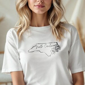 May include: White t-shirt featuring a black outline of the North Carolina state map with the words "North Carolina" and a floral design. The shirt is made of a soft material.