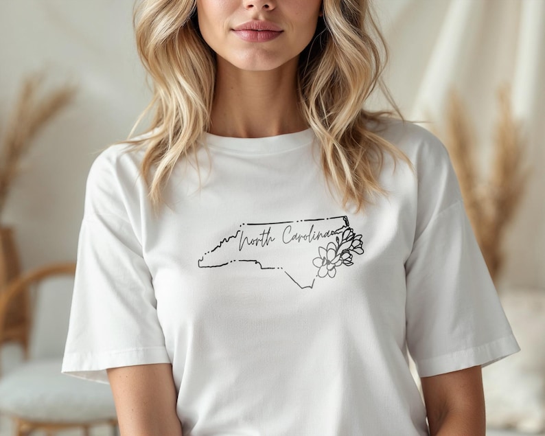 May include: White t-shirt featuring a black outline of the North Carolina state map with the words "North Carolina" and a floral design. The shirt is made of a soft material.