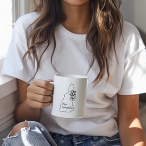 May include: White ceramic mug with a black outline of the state of New Hampshire and a floral design. The text "New Hampshire" is written in cursive below the state outline.