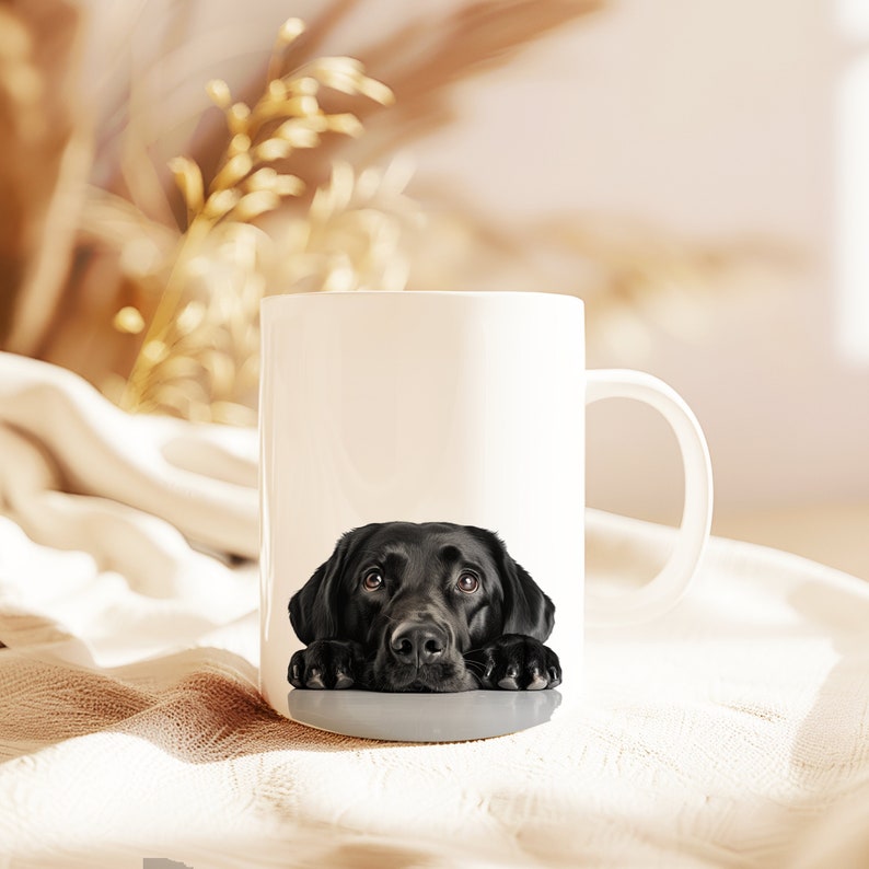 May include: A white ceramic mug with a black Labrador Retriever peeking over the rim. The dog has brown eyes and is looking directly at the viewer.