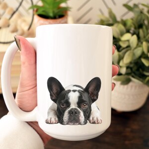 Op de afbeelding: Witte keramische mok met een zwart-wit Franse Bulldog ontwerp. De hond wordt liggend afgebeeld. De mok heeft een grote handgreep en wordt door een persoon vastgehouden. De mok is een standaard formaat.