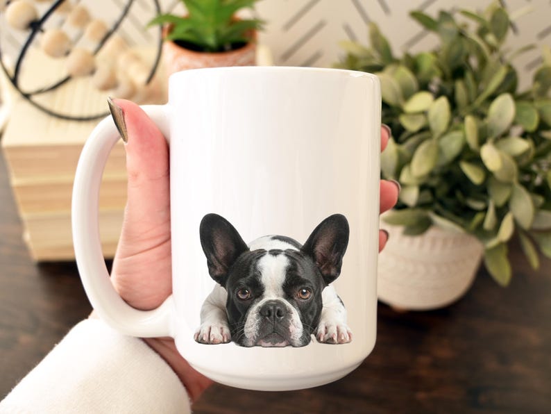 Op de afbeelding: Witte keramische mok met een zwart-wit Franse Bulldog ontwerp. De hond wordt liggend afgebeeld. De mok heeft een grote handgreep en wordt door een persoon vastgehouden. De mok is een standaard formaat.