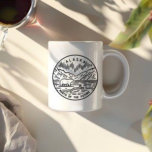 K&ouml;nnte beinhalten: Wei&szlig;e Keramiktasse mit einer schwarzen Linienzeichnung eines Eisb&auml;ren vor Bergen und der Aurora Borealis. Der Text "ALASKA NORTH OF THE FUTURE" ist auf die Tasse gedruckt.