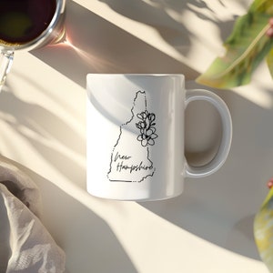 May include: White ceramic coffee mug with a black outline of the state of New Hampshire and a floral design. The text "New Hampshire" is written in cursive below the state outline.