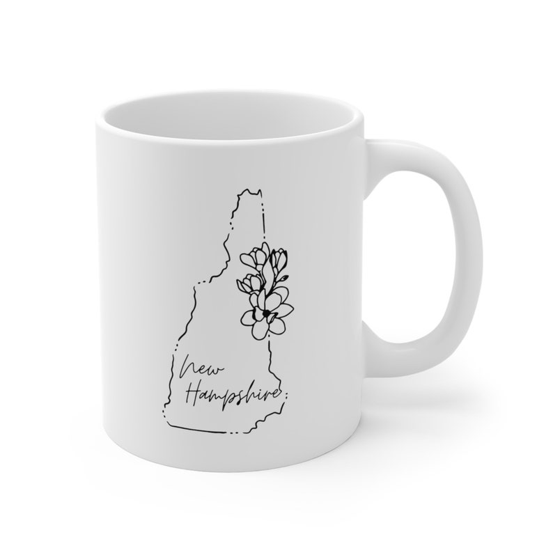May include: White ceramic coffee mug with a black line drawing of the state of New Hampshire with a cluster of flowers on the side. The text "New Hampshire" is written in cursive below the state outline.