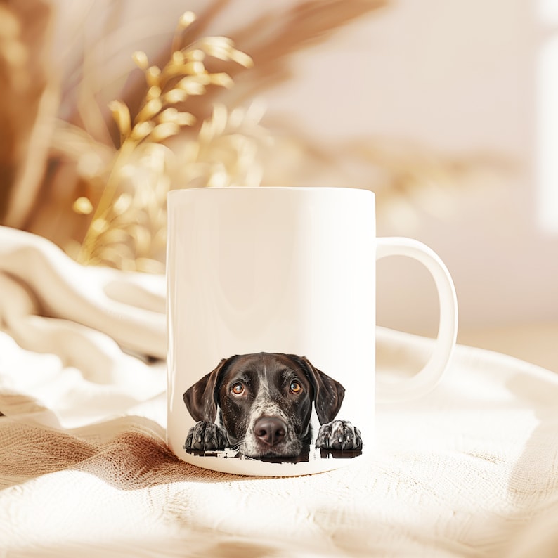 May include: White ceramic mug with a black and brown dog peeking over the rim. The dog has brown eyes and a spotted nose.