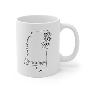Pode incluir: Caneca de cer&acirc;mica branca com um contorno preto do estado do Mississippi e um design floral. O texto "Mississippi" est&aacute; escrito em it&aacute;lico.