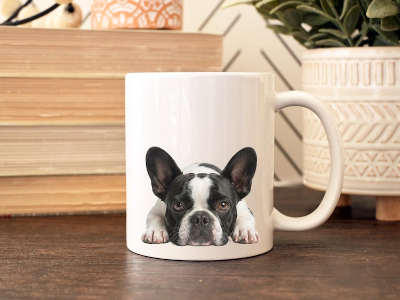 Op de afbeelding: Witte keramische mok met een zwart-wit Frans Bulldog ontwerp. De hond wordt liggend afgebeeld. De mok heeft een gebogen handvat en staat op een houten oppervlak.