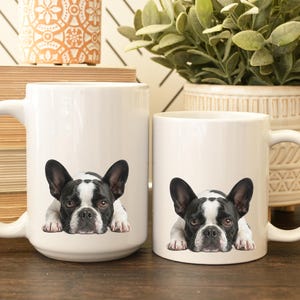 Op de afbeelding: Twee witte keramische mokken met een zwart-wit Franse Bulldog-ontwerp. De mokken, van verschillende afmetingen, hebben grote handvatten. De bulldog wordt liggend afgebeeld. De mokken staan op een houten oppervlak.