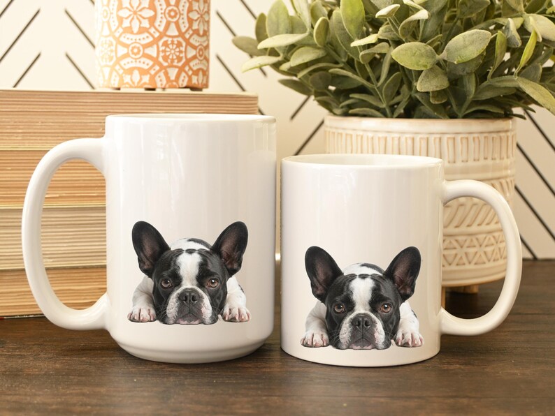 Op de afbeelding: Twee witte keramische mokken met een zwart-wit Franse Bulldog-ontwerp. De mokken, van verschillende afmetingen, hebben grote handvatten. De bulldog wordt liggend afgebeeld. De mokken staan op een houten oppervlak.