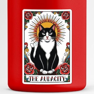 May include: A red water bottle with a sticker featuring a black and white cat illustration. The cat is surrounded by a golden halo, roses, and the text "THE AUDACITY".