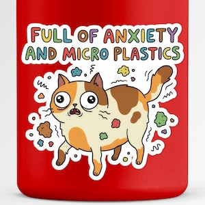May include: A red mug featuring a cartoon cat with a worried expression and the text "FULL OF ANXIETY AND MICRO PLASTICS" in colorful letters. The cat has brown, orange, and white fur and is surrounded by colorful specks.