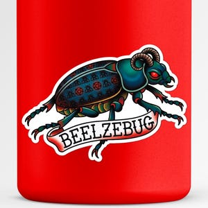 May include: A red tumbler featuring a detailed illustration of a beetle with horns and red eyes. The beetle has a blue and teal body with a decorative pattern. A banner below reads "BEELZEBUG".