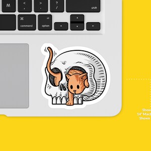 K&ouml;nnte beinhalten: Ein Aufkleber mit einem wei&szlig;en Sch&auml;del, aus dem ein kleines orangefarbenes K&auml;tzchen herausschaut. Der Aufkleber ist auf einem 14-Zoll-Macbook Pro-Laptop zu sehen.