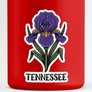 May include: A bright red cylindrical container featuring a sticker of a purple iris flower with green leaves and a yellow center. Below the flower, a white rectangular label with a black border displays the word TENNESSEE in black text.