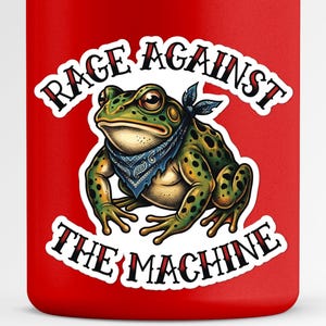 Puede incluir: Vaso rojo con una ilustración detallada de una rana verde con un pañuelo azul. El texto "RAGE AGAINST THE MACHINE" se muestra en una fuente estilizada encima y debajo de la rana. El diseño es una declaración audaz.