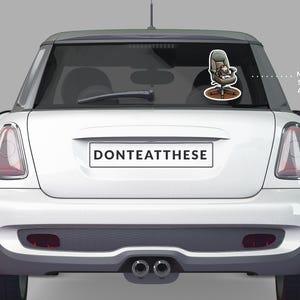 Puede incluir: Un Mini Cooper blanco con una pegatina en la luneta trasera que muestra un gato de dibujos animados en una silla de oficina. La pegatina mide aproximadamente 28 cm. La matr&iacute;cula dice "DONTEATTHESE".