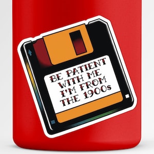 Puede incluir: Una pegatina con un diseño de disquete retro con el texto "BE PATIENT WITH ME I'M FROM THE 1900s". La pegatina tiene una combinación de colores negro, naranja y rojo y está colocada sobre una superficie roja.