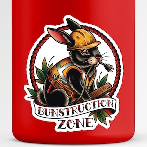 May include: A red tumbler featuring a sticker with a black rabbit wearing a yellow hard hat and construction vest. The rabbit holds a plug in its mouth, with the words "Bunstruction Zone" on a banner.