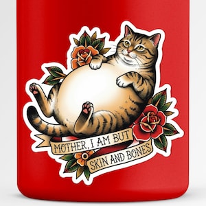 May include: A red tumbler featuring a cartoon cat tattoo design. The cat is brown and tan, with a large belly, surrounded by roses and a banner that reads "MOTHER, I AM BUT SKIN AND BONES."