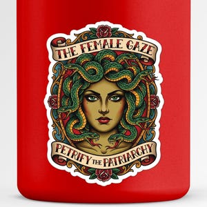 Può includere: Borraccia rossa con un'illustrazione di Medusa, con serpenti verdi al posto dei capelli, e il testo "THE FEMALE GAZE" e "PETRIFY THE PATRIARCHY" su uno striscione.