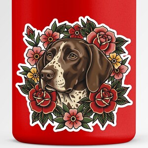 May include: A red tumbler featuring a German Shorthaired Pointer dog illustration surrounded by red roses, pink and yellow flowers, and green leaves. The dog has brown and white markings. The design is a tattoo-style graphic.
